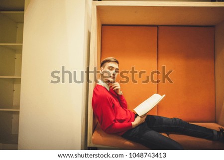 Young male student sitting on windowsill in library and reading book