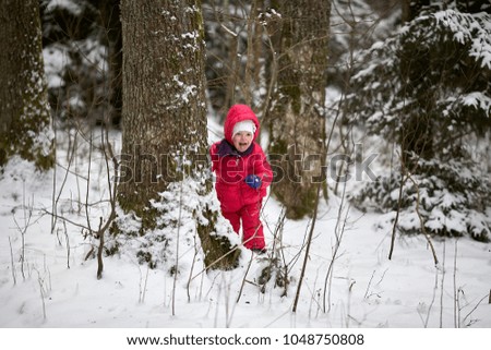 cute little baby girl child with red jacket and pants is playing in the forest park in winter.