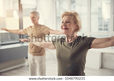 Waist up portrait of old jolly lady with arms straightened out aside. Male pensioner in same posture standing on background. Focus on woman Royalty-Free Stock Photo #1048790231