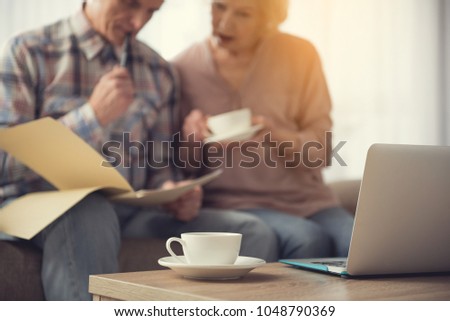 Focus on mug and laptop on table. Elderly couple focused on documents on background Royalty-Free Stock Photo #1048790369