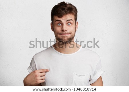 Funny bearded male presses lips, stares at camera, points at blank casual t shirt, isolated over white studio background. Bugged eyed brunet young man with bristle poses in studio. Advertisment