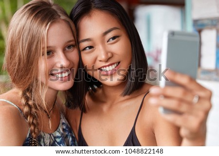 Happy multiethnic girlfriends exciting with leisure, hold modern cell phone for making selfie, pose in canera with pleasant smiles, meet in restaurant. Chinsese and European women take picture