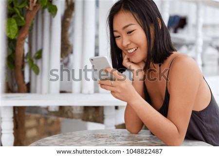 Positive young woman with narrow eyes and bobbed hairstyle share media in social networks or installs new app on mobile phone, sits against cafe interior. Asian female reads positive news online