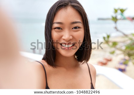 Pretty young Asian woman takes photo on camera og unrecognizable gadget, poses against sea or ocean horizon, takes picture during free time, enjoys good rest. Outdoor shot of cute Chinese girl