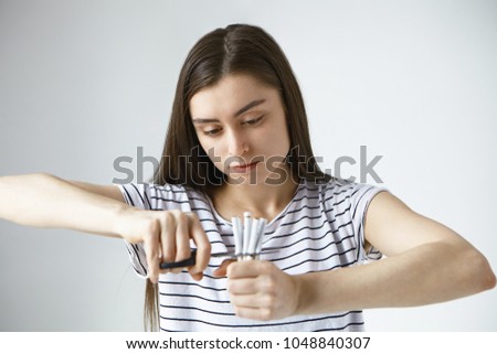 Picture of decisive strong willed young dark haired woman dressed casually cutting bunch of cigarette in halves with scissors, having determined look, quitting smoking. People and bad habits concept