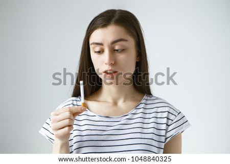 Picture of worried perplexed young brunette female wearing casual t-shirt holding cigarette she found in her husband's bag, looking at it with disappointment. People, bad habits and addiction concept
