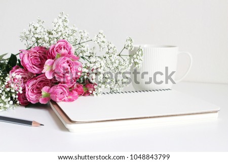 Styled stock photo. Closeup of wedding bouquet made of pink roses and baby's breath, Gypsophila flowers lying on white table. Feminine still life, blank notebook, pencil and white cup of coffee. 