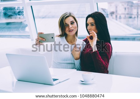 Two attractive bloggers making selfie on modern telephone while sitting in stylish coffee shop with digital laptop computer.Positive hipster girls taking photos on cellular in university interior