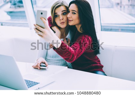 Cheerful internet users enjoying recreation time while making funny selfie photos on front camera of cellphone.Brunette female holding smartphone in hand and taking positive pictures with best friend