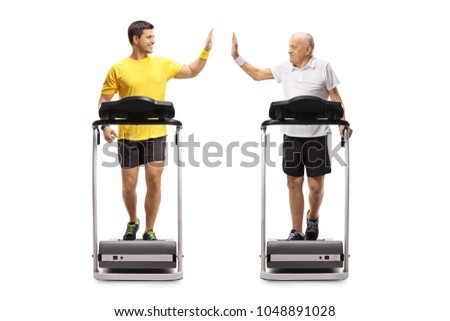 Full length profile shot of a young man and a senior walking on treadmills and high-fiving each other isolated on white background