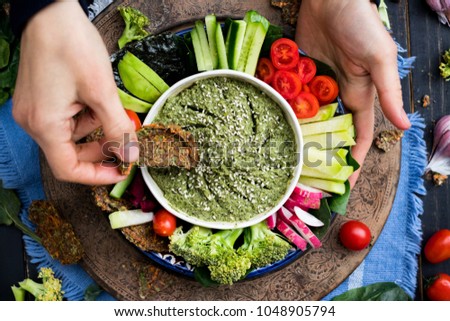 Woman hands try the dipping raw vegan flax bread into Italian pesto sauce in vegetarian healthy buddha bowl with vegetables - tomatoes, cucumbers, spinach, radish, daikon, broccoli. Vegetarian food.