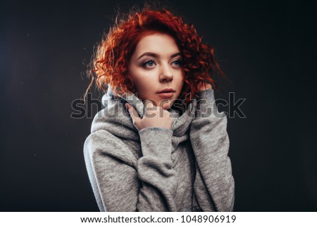 young cute redhair girl with blue eyes wrapped himself in a gray hoodie large portrait in studio on the black background