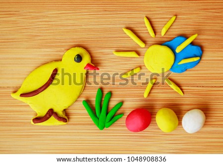 Funny spring Easter image made of plasticine / Yellow chicken, green grass, colorful eggs - red, yellow and white, golden sun shining over blue cloud on wooden background, warm colors