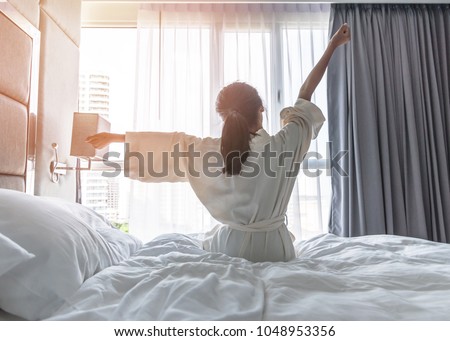 Easy lifestyle Asian woman waking up from good sleep in weekend morning taking some rest, relaxing in comfort bedroom at hotel window, having happy lazy day enjoying work-life quality balance concept Royalty-Free Stock Photo #1048953356