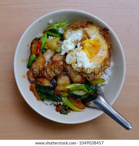 White rice cover by fried green vegetable, red chilli and crispy pork or "stir fry kai land and crispy pork" with fried egg and stainless spoon in a blow. on light brown background. Square top view.  