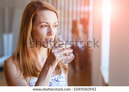 Portrait of good morning with glass of water holding by pretty blond middle age woman. Concept of drinking fresh. Sun glare.