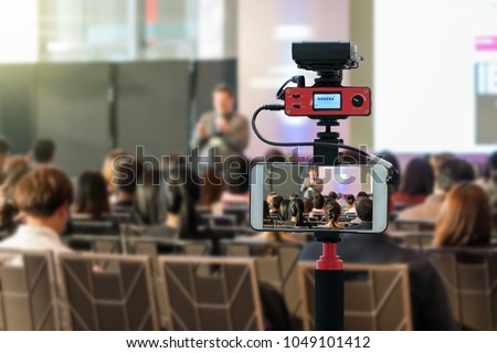 Closeup smart mobile phone taking Live over Speakers on the stage with Rear view of Audience in the conference hall or seminar meeting, technology live streaming and broadcast concept