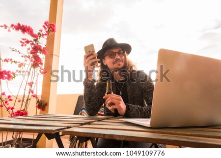 trendy man with hat and eyeglasses drink a beer while use the phone to ommunicate with friends and people. laptop on the table outdoor terrace to work free