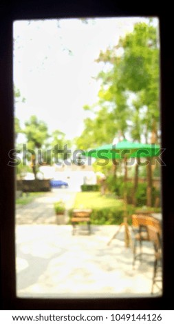 Look outside through window frame. It is very sunny and in garden has  wooden chair, green umbrella. This picture is blur. 