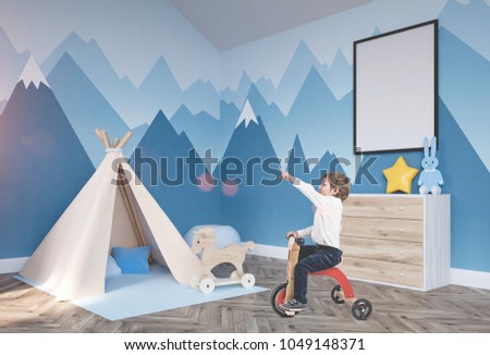 Cute little boy in a white shirt and dark blue jeans is riding a tricycle and showing with his finger. A baby boy room background with a poster mock up and a tent
