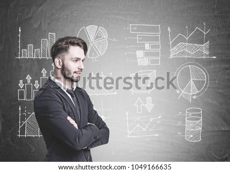 Side view of a pensive young businessman with a beard. He is wearing a suit and a tie and standing with crossed arms. Graphs and diagrams drawn on a blackboard