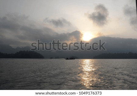 sunlight reflects from water in lake at sunset surrounding 
the sailing ship, Small island of trees and low clouds.

