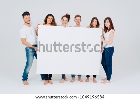 Group of casual barefoot friends in jeans with a blank white signboard in front of them standing in a line smiling at the camera