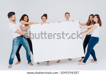 Playful group of casual barefoot friends tugging on a large white blank rectangular sign with copy space on a white studio background
