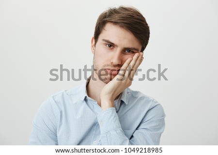Indoor shot of upset irritated caucasian guy leaning on palm and sighing at camera, being bored and fed up of annoying meeting in office, wanting to go home, tired of wasting time over gray wall Royalty-Free Stock Photo #1049218985