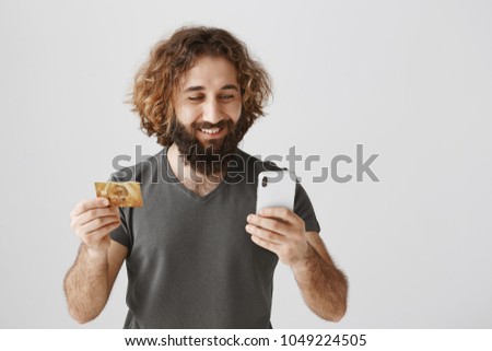 Glad to have deal with great bank. Studio shot of pleased eastern client holding credit card and smartphone, looking at screen with smile, satisfied with fast online banking system over gray wall