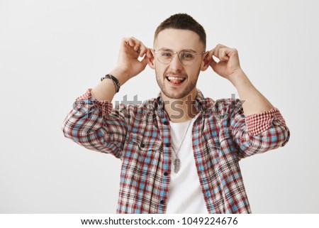 Guy with elf ears thinks they add charm to appearance. Portrait of good-looking funny man in eyeglasses holding ears with hands and showing tongue, smiling and standing over gray background