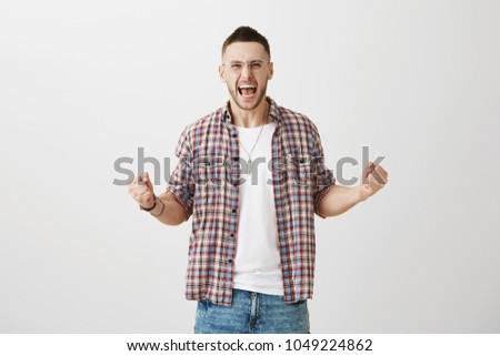 Emotive adult in glasses and plaid shirt shouting fearfully and squeezing raised fists, being very excited of his football team victory, standing over gray background. Guy feels sweet victory taste