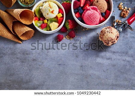 Summer ice cream flavors with fresh fruit salad, assorted berries, nuts, sugar cones and a scoop as a border on textured slate with copy space