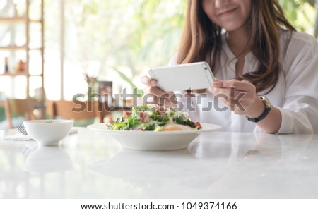 Closeup image of an asian woman using smartphone to taking photo of salad in a white plate on table in cafe