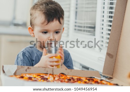 the child in the kitchen drinks orange juice and there is a large pizza
