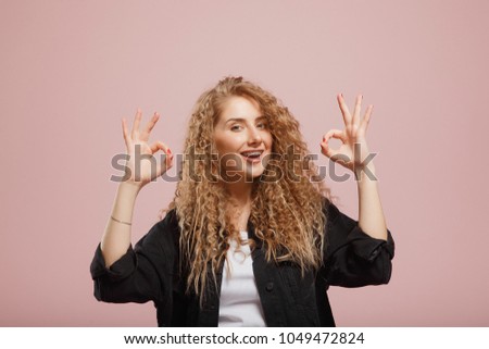Glad attractive woman curly shows okay as expresses approval, gestures in studio against pink background, has cheerful expression. Photo of beautiful female appealing appearance, being optimstic