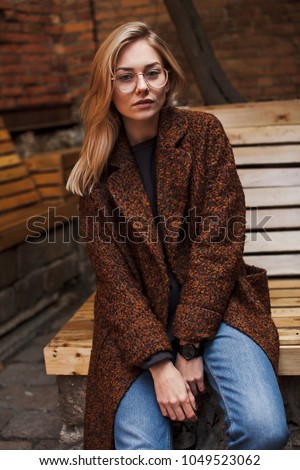 Young beautiful woman sits at wooden bench wearing terracotta coat and glasses. Hipster girl with blonde hair posing on street