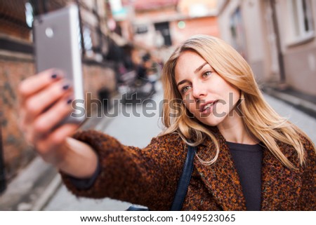 Blonde girl in terracotta coat wearing casual outfit making selfie portrait with her phone outdoors having fun and fooling