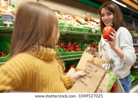Back view portrait of little girl helping mom grocery shopping in supermarket holding list with foods to buy while choosing vegetables, copy space