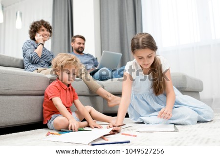 Ordinary day of large family: curly little boy and his elder sister sitting on carpet and drawing pictures with pencils while their pretty mother talking on phone and their bearded dad using laptop