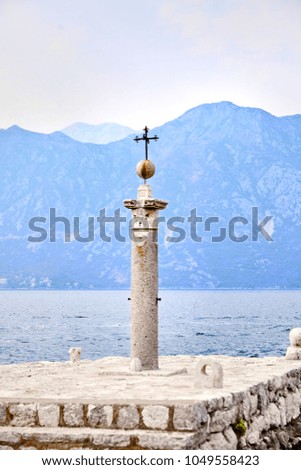 The stone column with the iron cross on the top of ball on Our lady of the Rocks Island, near Perast, the Adriatic Sea, Montenegro. On mountains background