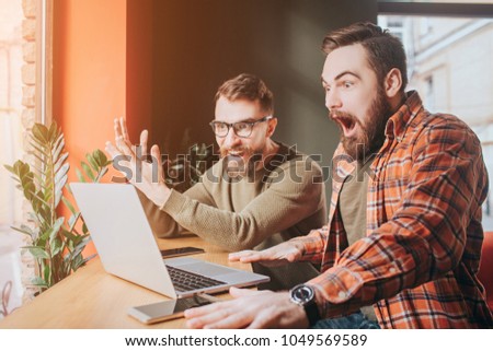 Very emotional picture of guys looking to the laptop's screen. One of them is very amazed and opened his mouth very wide while another is just looking to the screen.