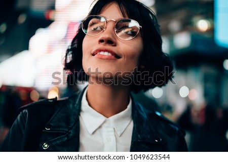 Fascinated pretty hipster girl in stylish eyeglasses amazed with night New York city walk in urban setting. Closeup excited stylish young woman standing in night city against blurred bokeh lights Royalty-Free Stock Photo #1049623544