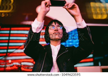 Amazed brunette hipster girl in eyewear enjoying night walk in New York city and making photos on smartphone for publication in blog.Excited woman in stylish wear shooting video with neon illumination