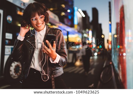 Cheerful pretty young woman in cool eyeglasses and trendy wear walking on metropolis street with night lights enjoying online playlist songs in earphones and reading sms with good news on smartphone Royalty-Free Stock Photo #1049623625