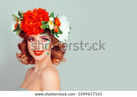 Beautiful romantic young woman in a wreath of flowers red white peony floral headband looking to the side red green flower painting on face posing on a light green studio wall background