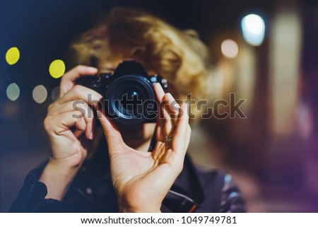 Hipster hiker tourist making photo, holding in hands camera on background of evening city, blogger view in holiday, photographer girl enjoying night street in trip, traveler relax lifestyle concept