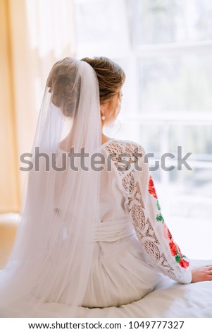 bride in a white dress with lace and embroidery with a veil sits on the bed and looks out the window