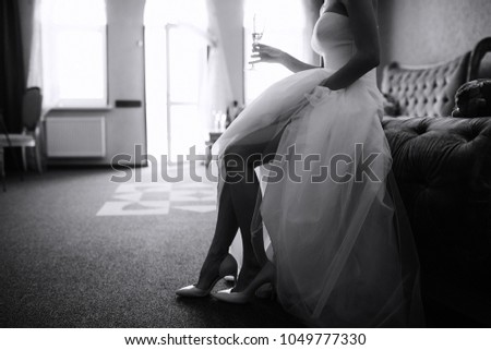 bride wearing a wedding dress sits on the couch and holds the glass with her other hand slightly lifting her skirt against the background of a large window