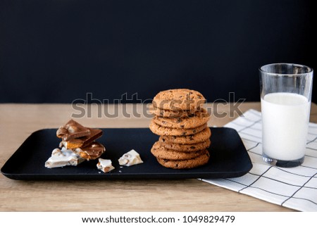 black plate on which lies a cookie and black with white chocolate with nuts near which stands a glass of milk on a wooden table with a white napkin on a black background